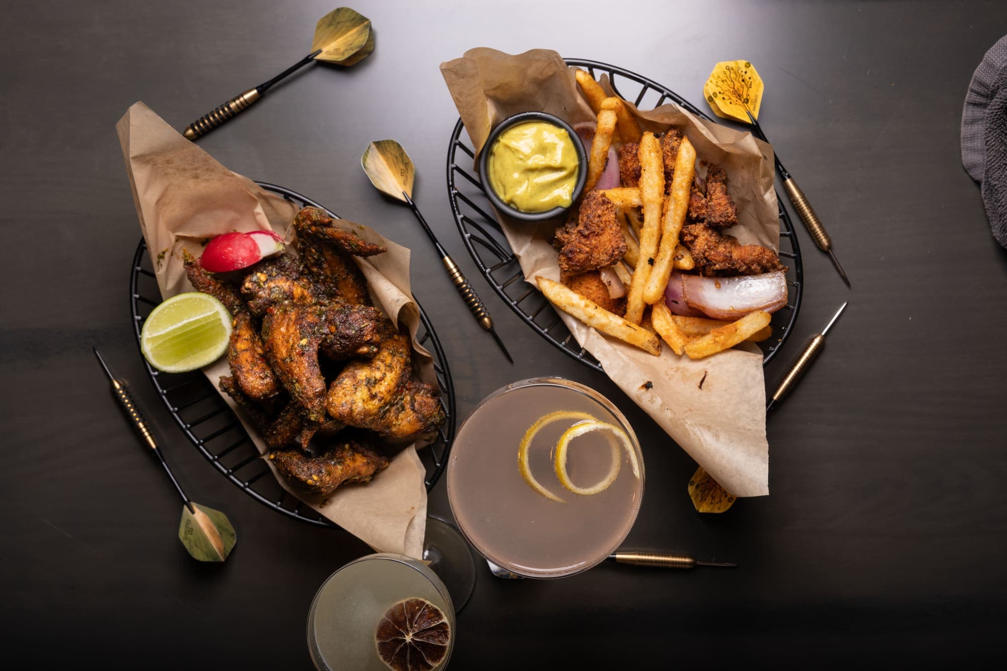 Chicken wings and fried chicken tenders with fries served alongside cocktails and darts